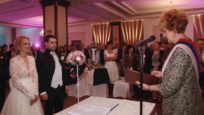 Mladenci stoje ispred matičara tokom građanskog venčanja u svečanoj sali sa gostima.