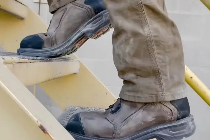 Radnik u zaštitnim radnim cipelama hoda po metalnim stepenicama u industrijskom prostoru.
